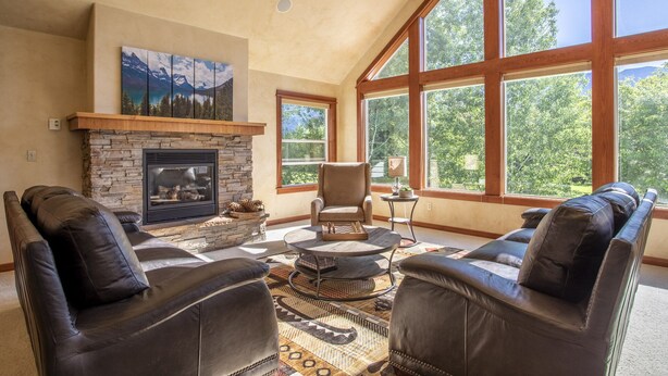 A spacious living room with a stone fireplace, leather sofas, and large windows offering a view of greenery. - Gather your family in Montana for outdoor fun and memories. (Kalispell)