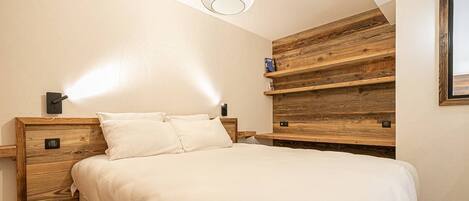 Quarto com cabeceira de madeira, cama com roupa de cama branca e estante embutida com acabamento rústico.