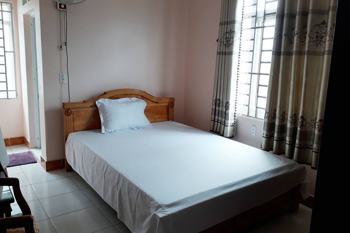 Une chambre à coucher avec une tête de lit en bois, un seul oreiller blanc, un lit avec des draps blancs, un ventilateur sur pied et une fenêtre avec des rideaux.