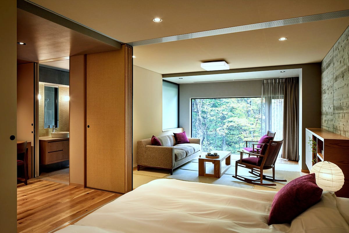 A modern bedroom with a large bed, a sofa, a rocking chair, a small table, and a view of trees through a large window.