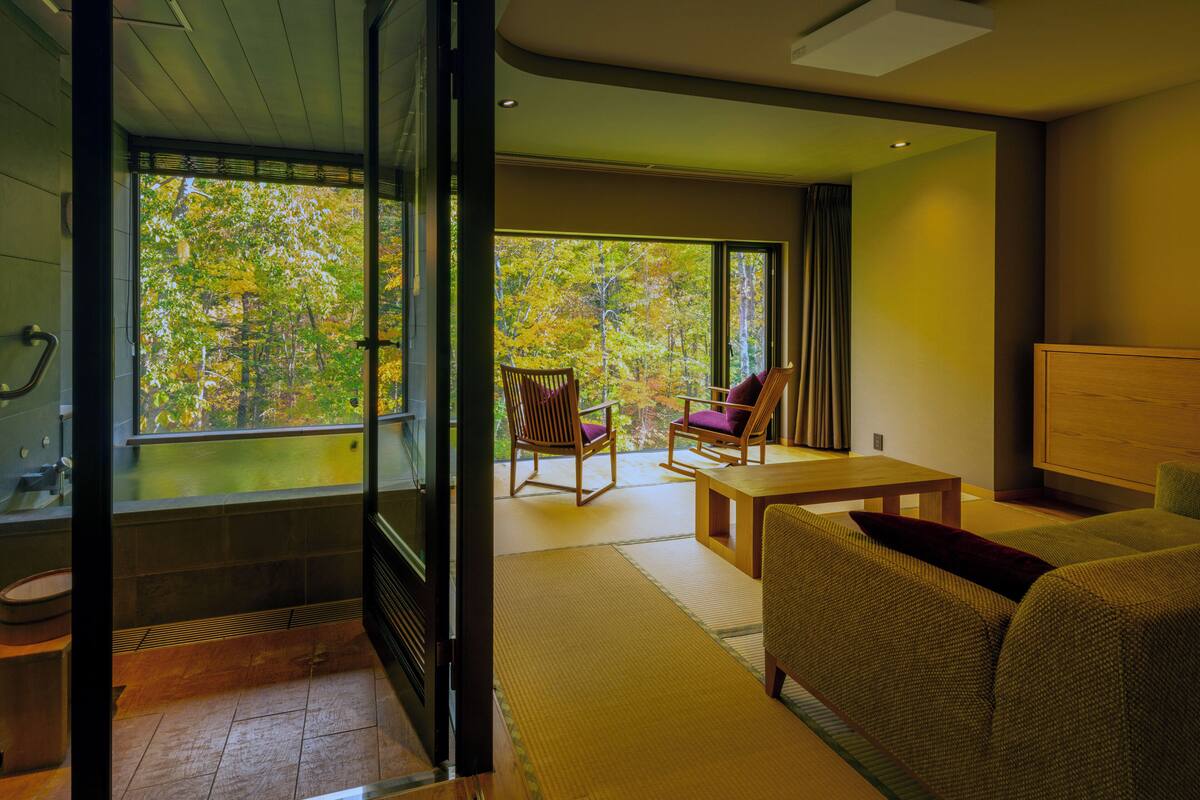 A modern hotel room with a large sliding glass door leading to a balcony, a wooden chair, a small table, and a sofa.