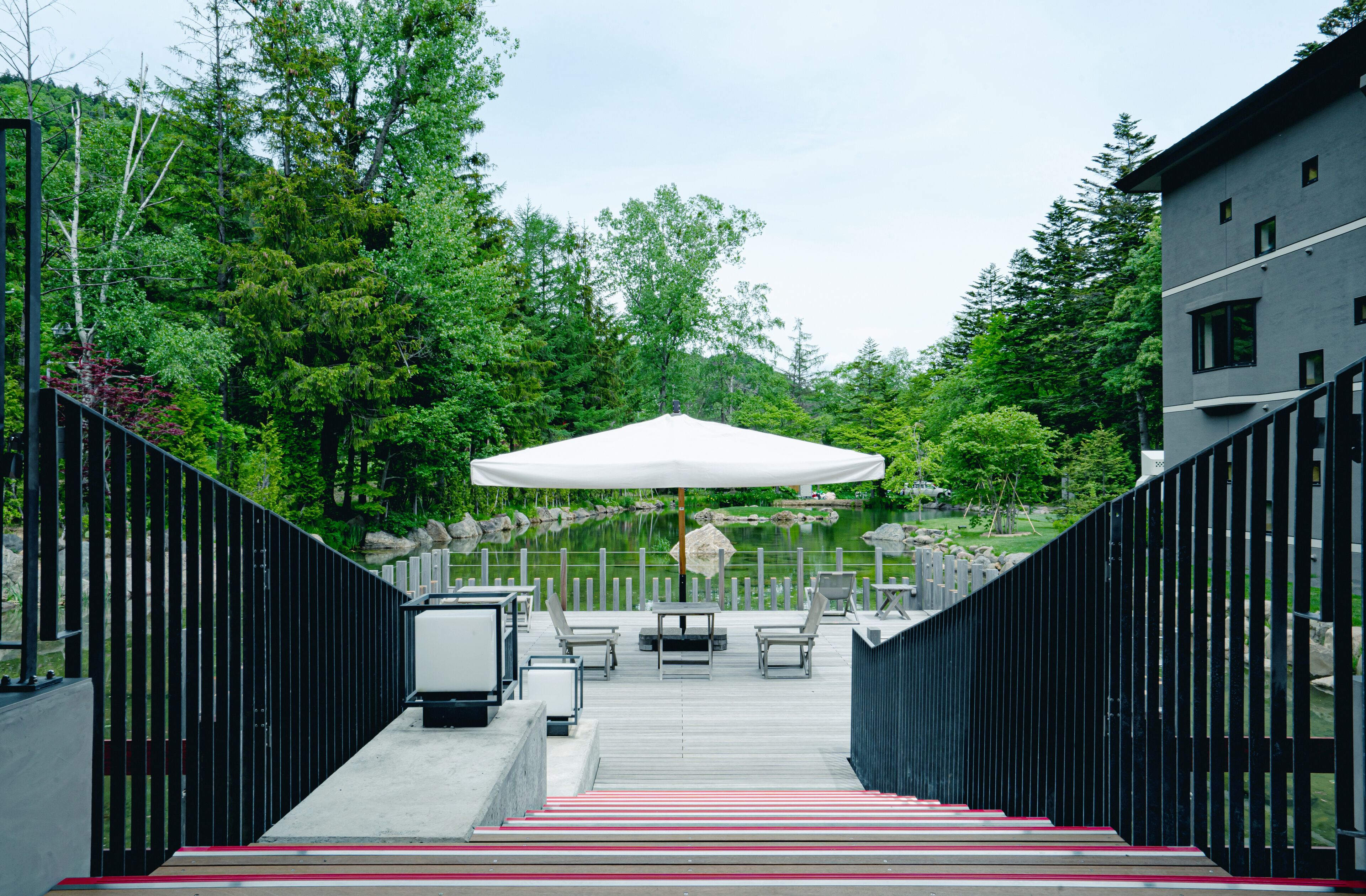 A wooden deck with outdoor seating, a white umbrella, and a view of a pond surrounded by trees.