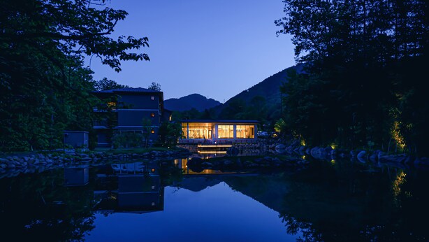 A lakeside building with a reflection in the water, surrounded by trees and hills. - Okujozankeionsen Kashogyoen (Sapporo)