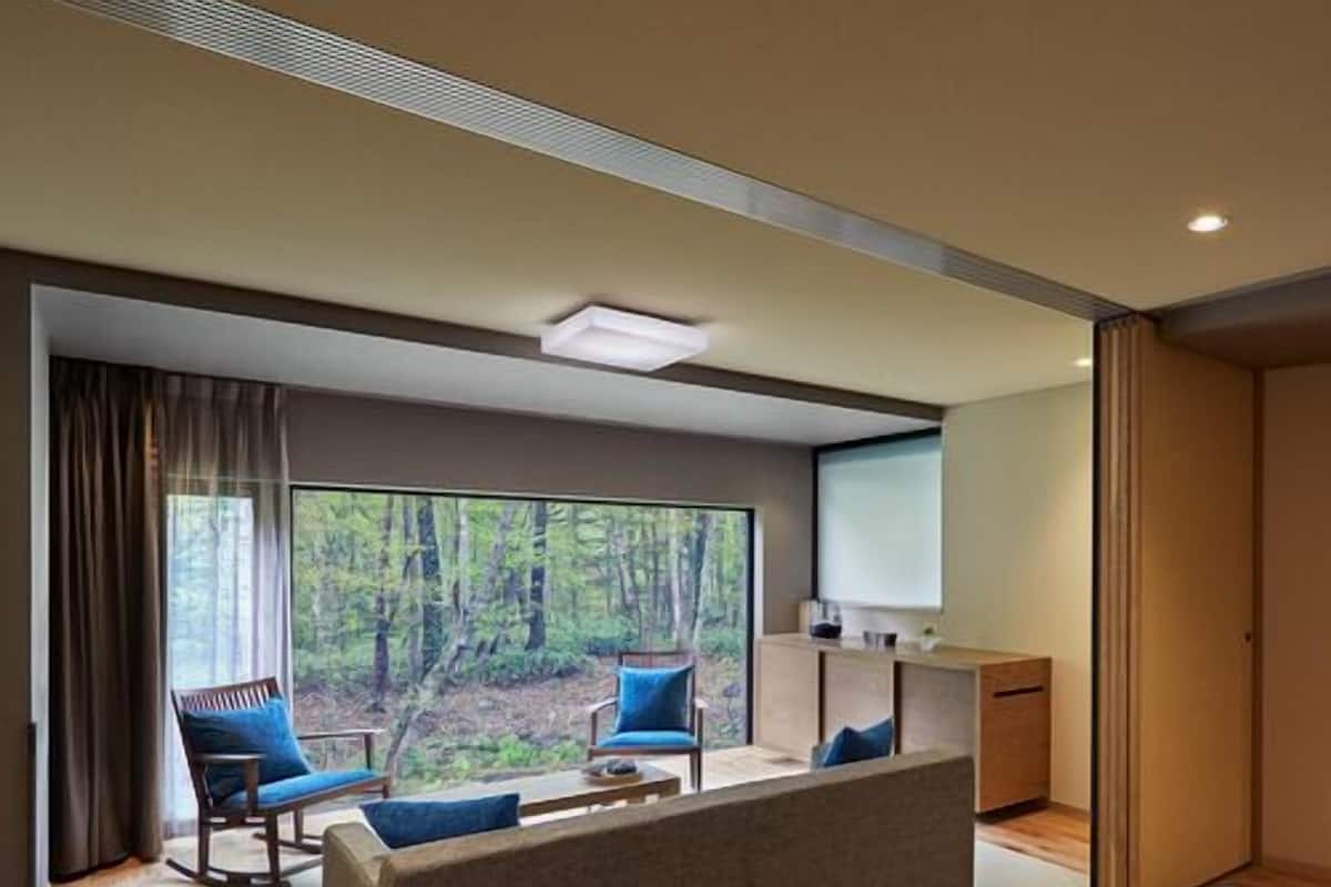 A modern hotel room with a large bed, a sofa, two armchairs, a TV, and a view of a forest.