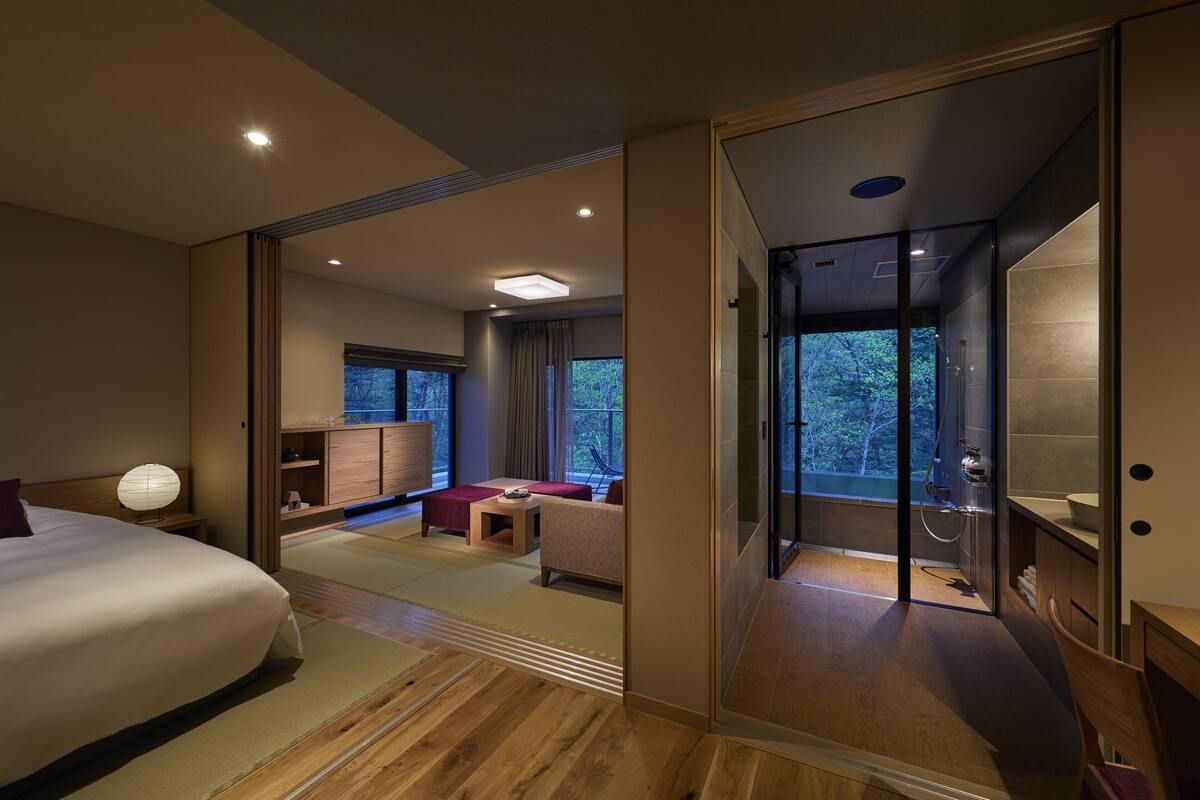 A modern hotel room with a large bed, a seating area with a sofa and a small table, a glass-enclosed shower area, and a view of the outdoors.