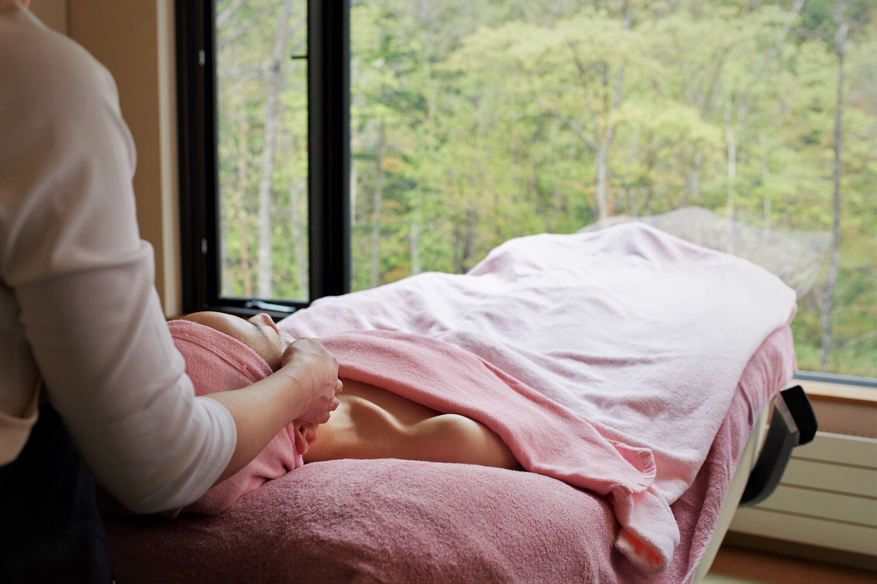 A person lying on a bed with a pink blanket, in a room with a window overlooking a forest.
