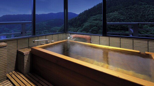 A hot tub with a wooden frame and a view of a forested mountain range through large windows. - bankokuya (Tsuruoka)
