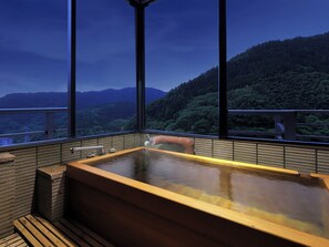 Un jacuzzi con estructura de madera y vistas a una cadena montañosa boscosa a través de amplios ventanales.
