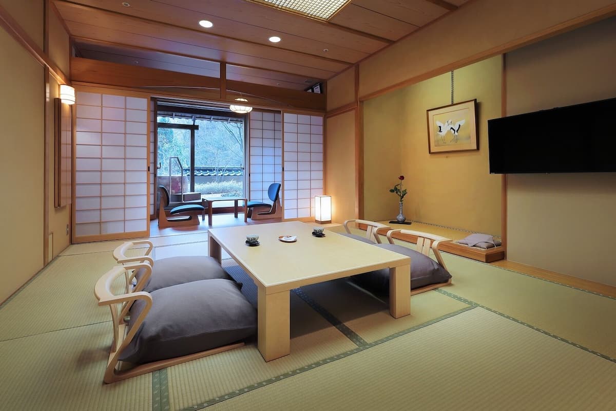 Ein Raum mit Tatami-Matten, ausgestattet mit einem niedrigen Tisch, Kissen, einem Fernseher und einer Schiebetür, die auf einen Balkon führt.