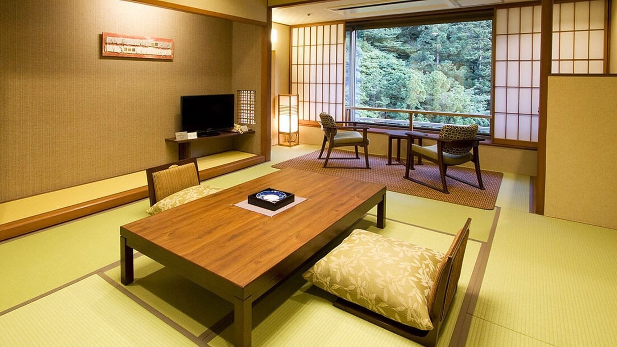 Una habitación japonesa tradicional con suelo de tatami, una mesa baja con cojines, un televisor y vistas a la vegetación a través de biombos shoji.