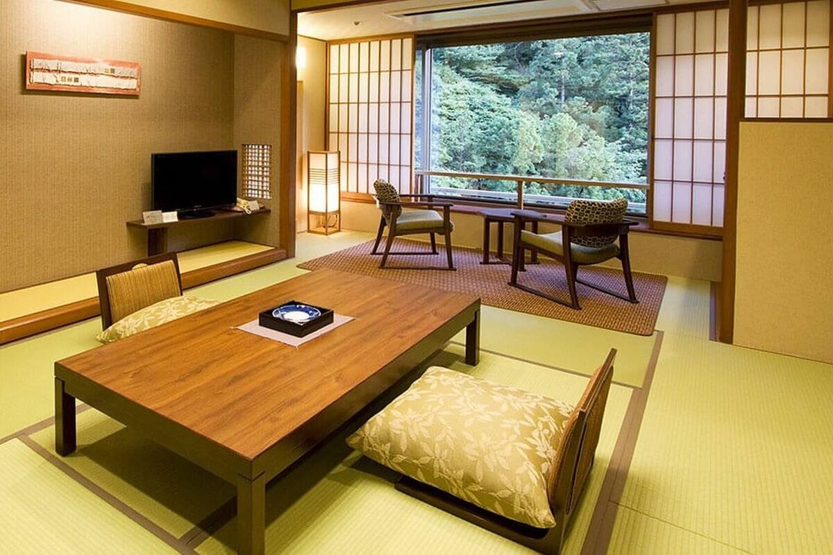 A traditional Japanese room with tatami flooring, a low table with cushions, a television, and a view of greenery through shoji screens.