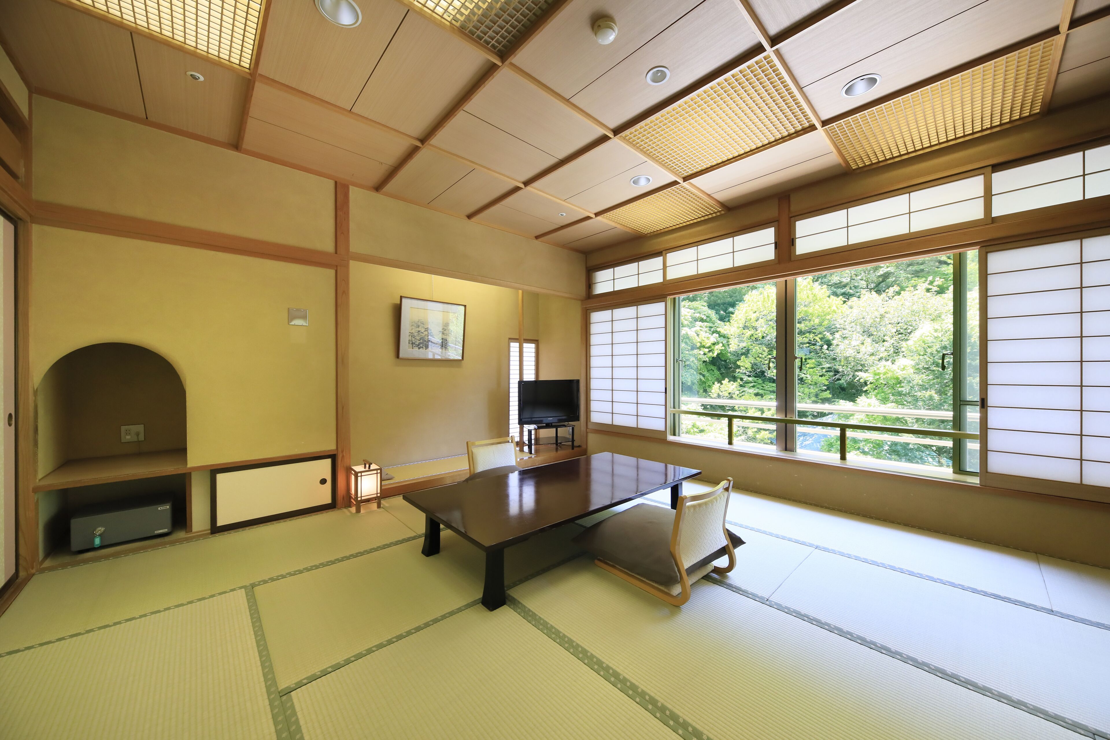 Ein traditioneller japanischer Raum mit Tatami-Böden, einem niedrigen Tisch, einem Fernseher und großen Fenstern mit Blick auf Grünflächen.