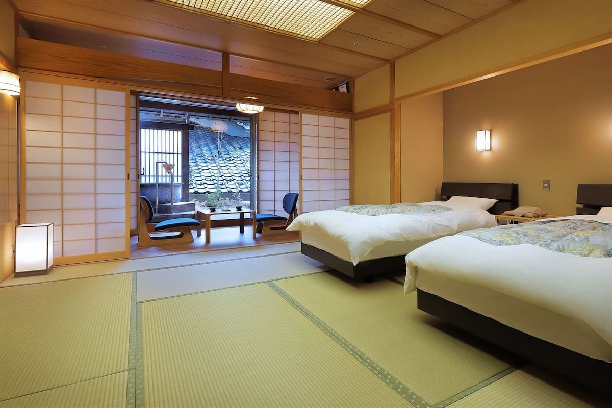 A traditional Japanese room with tatami flooring, two beds, a table with chairs, and a sliding door leading to a balcony.