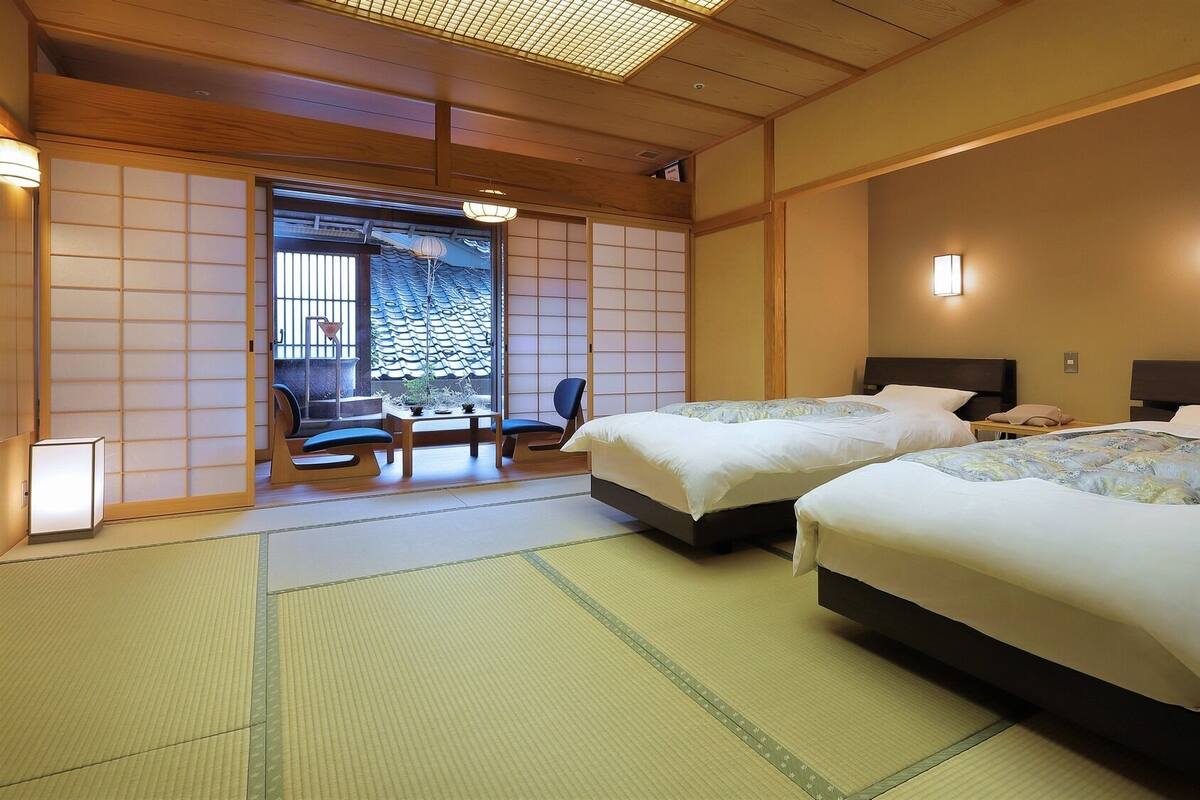 Una habitación tradicional japonesa con suelo de tatami, dos camas, una mesa con sillas y una puerta corrediza que da a un balcón.