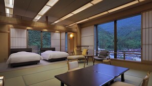 A hotel room with two beds, a dining table, chairs, and large windows offering a view of a forested area. - bankokuya (Tsuruoka)