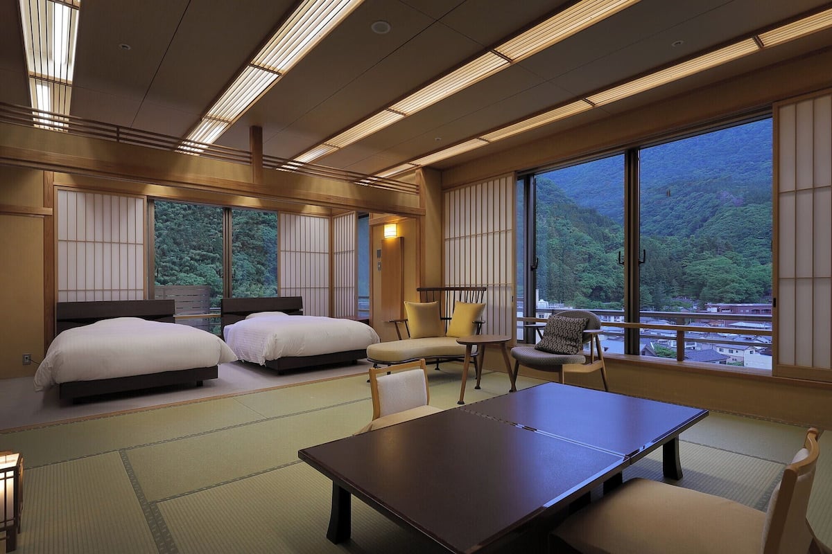 A hotel room with two beds, a dining table, chairs, and large windows offering a view of a forested area.