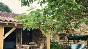 A rustic stone building with a wooden pergola, a round wooden table with chairs, and a grill area.