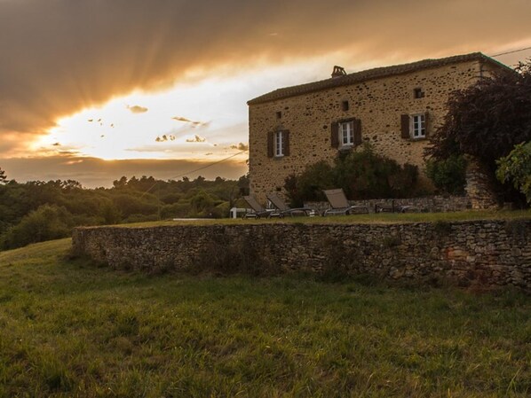 Un bâtiment en pierre au style rustique, avec un mur de pierre en premier plan et une zone herbeuse agrémentée de quelques chaises longues.