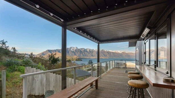 A wooden deck with a bench and bar stools overlooking a mountainous landscape and a lake.