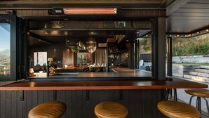 A modern bar area with a wooden counter, three stools, and a view of the outdoor landscape.