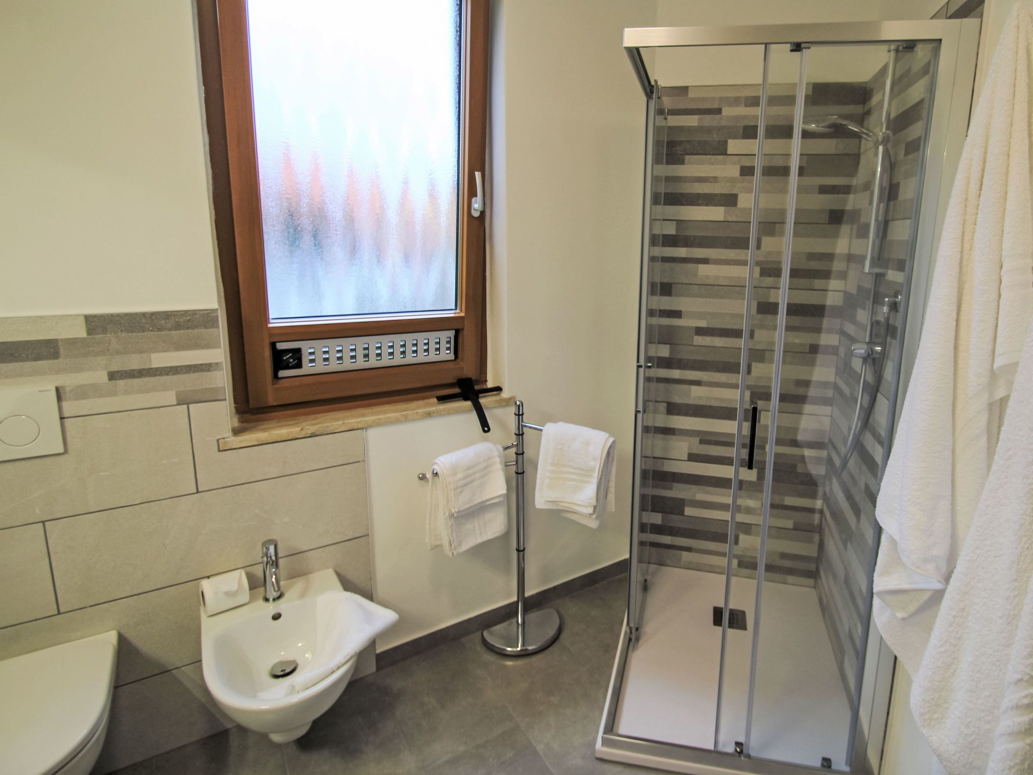 A modern bathroom with a glass shower enclosure, a bidet, and a window with frosted glass.