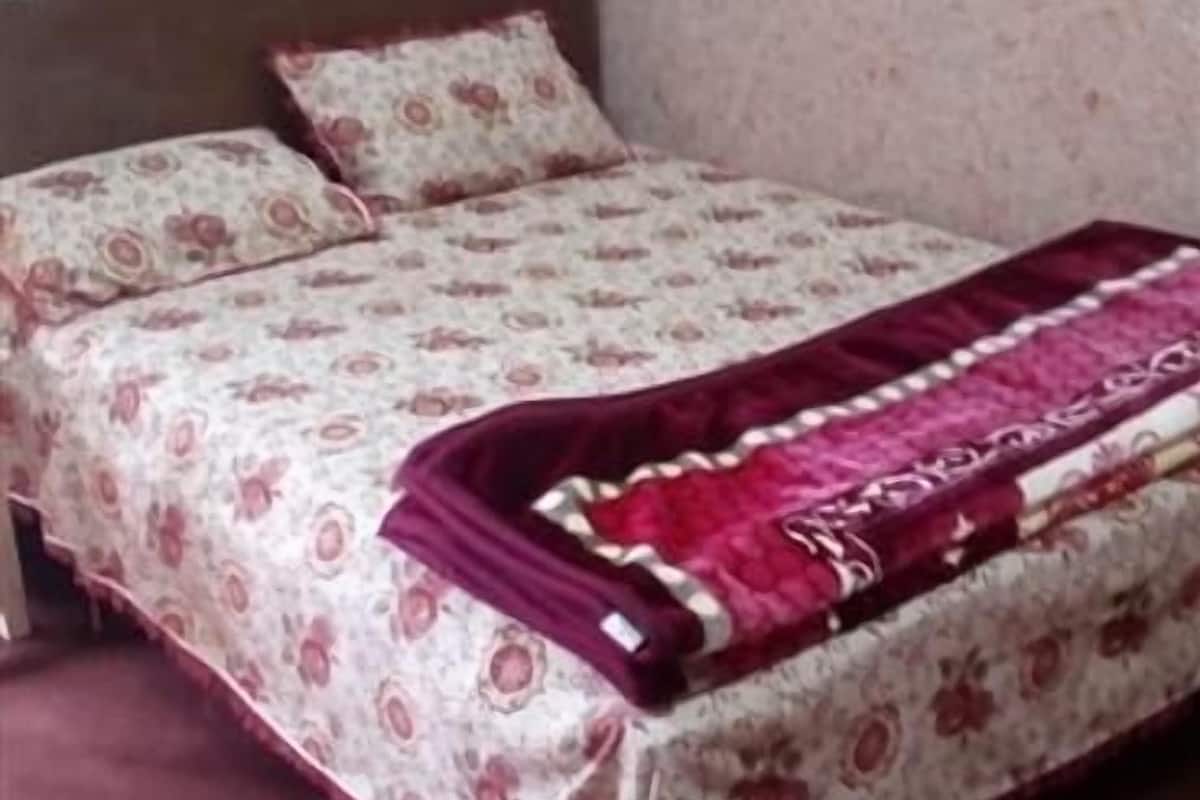 Uma cama com colcha floral e manta vermelha, cabeceira de madeira e cortinas estampadas.