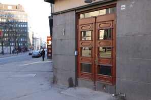 Un bâtiment doté d’une grande porte en bois, d’une boîte aux lettres bien visible et situé dans une rue où l’on croise des piétons et des voitures.