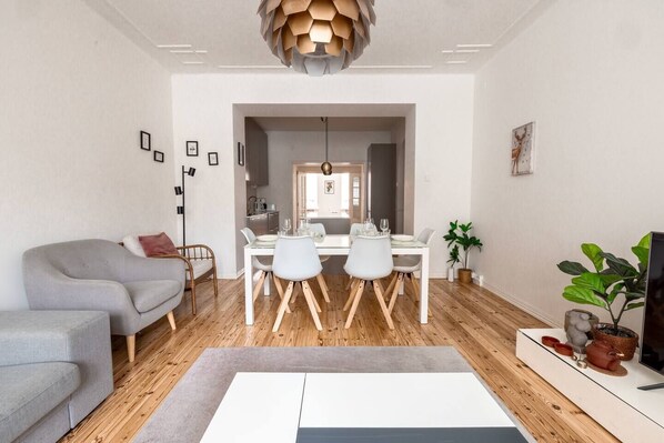 A modern living room with a grey sofa, a dining table with chairs, a TV, and a potted plant.
