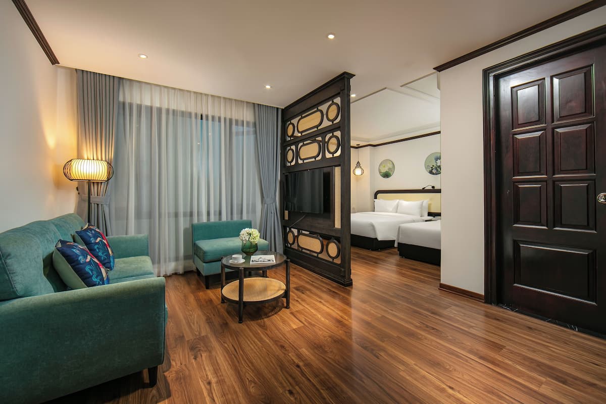 A hotel room with a green sofa, a small round table with a vase, a bed with white bedding, and a dark wooden door.