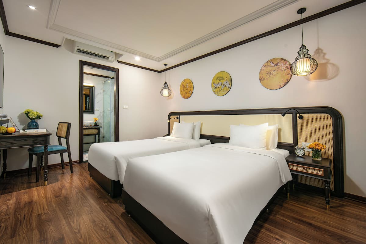 A hotel room with two beds, a desk with a chair, a small table with a vase of flowers, and wall art.