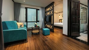 Una habitación de hotel moderna con un sofá color turquesa, una pequeña mesa redonda con un florero, un otoman azul, un ventanal con cortinas, una cama con cabecera negra y una puerta que da a un baño.