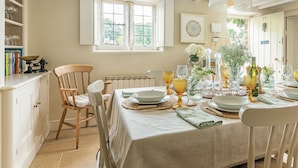 A dining area with a table set for four, surrounded by wooden chairs. A window provides natural light, and a bookshelf with books and decorative items is visible to the left. - Four bed Cotswold holiday cottage - Greenview Cottage (Little Barrington, near Burford)