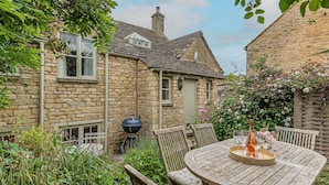 A stone house with a wooden table and chairs, surrounded by lush greenery and flowers. - Four bed Cotswold holiday cottage - Greenview Cottage (Little Barrington, near Burford)