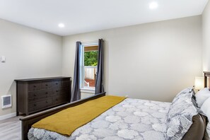 Une chambre à coucher avec un lit, une commode, une fenêtre avec des rideaux et une vue sur la verdure à l’extérieur.