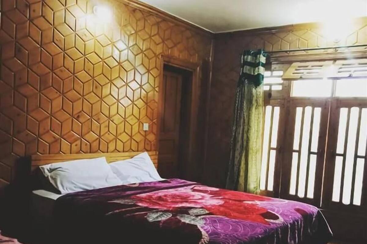 A bedroom with a wooden hexagonal wall pattern, a bed with a colorful blanket, and a window with vertical blinds.