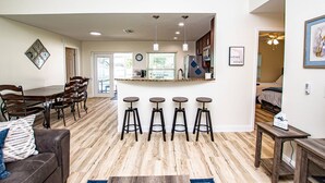 Moderni keittiö, jossa on baarituoleja, ruokailutila puupöydällä ja -tuoleilla sekä olohuone, jossa on sohva ja sohvapöytä.