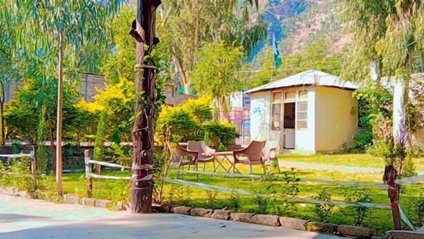 A small, single-story building with a flat roof, surrounded by greenery and trees. There are outdoor chairs and a table visible in front of the building. - Indus Hotel Shangla (Besham City)