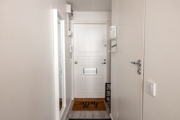 A hallway with a white door, a doormat with the word "welcome," and a small shelf with a telephone and a basket.