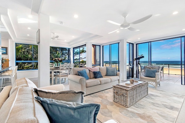 A spacious living area with a balcony overlooking the ocean, large windows, and a ceiling fan.