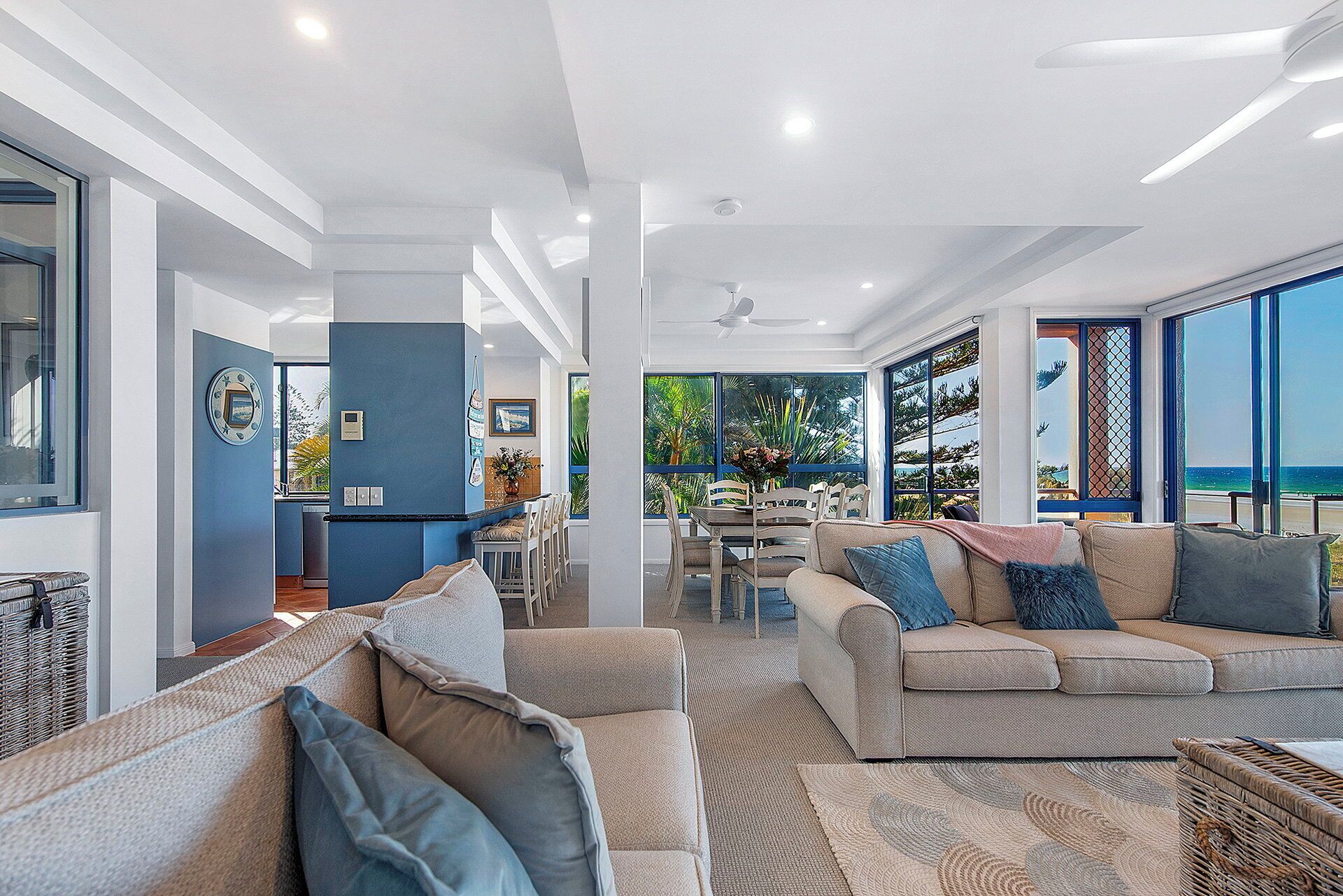 A modern living room with a sofa, a dining area, and large windows offering a view of the outdoors.
