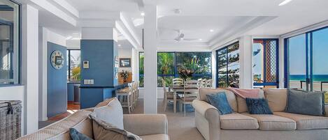 A modern living room with a sofa, a dining area, and large windows offering a view of the outdoors.