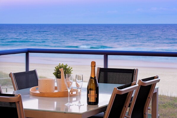 A table with a bottle of Chandon Brut wine, two glasses, a woven basket, and a small plant, set against a beach view.