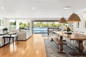 Amplia sala de estar con vista a la piscina, suelo de madera y zona de comedor con una mesa larga y bancos.