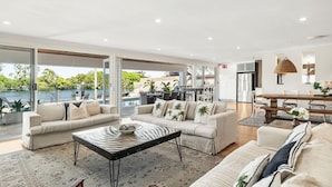A spacious living area with a large window overlooking a pool and greenery, a dining area with a wooden table, and a kitchen in the background. - Surfers Paradise Escape (Huge Heated Pool) (Gold Coast)