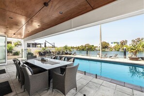 Un área de piscina exterior moderna con una mesa y sillas para comer, rodeada de macetas con plantas y vistas a un cuerpo de agua.