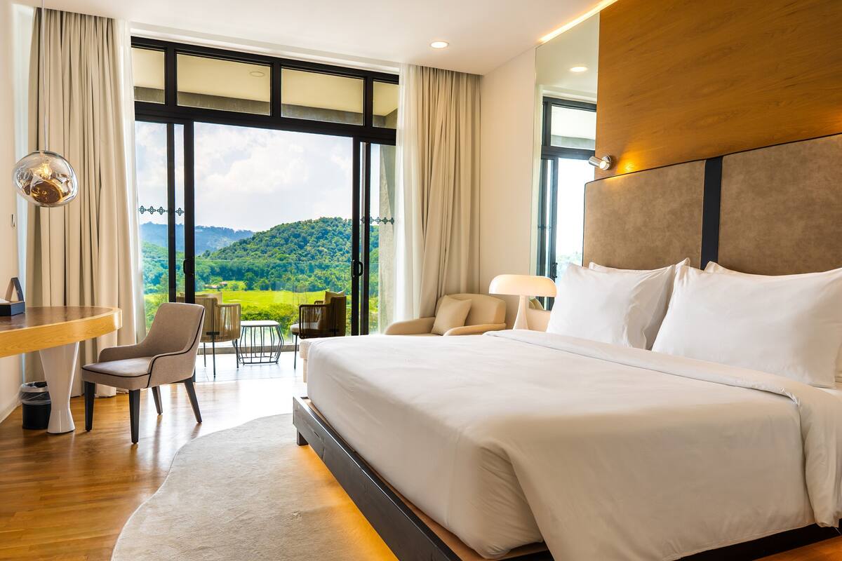 A hotel room with a large bed, a desk, and a view of a green landscape through large windows.