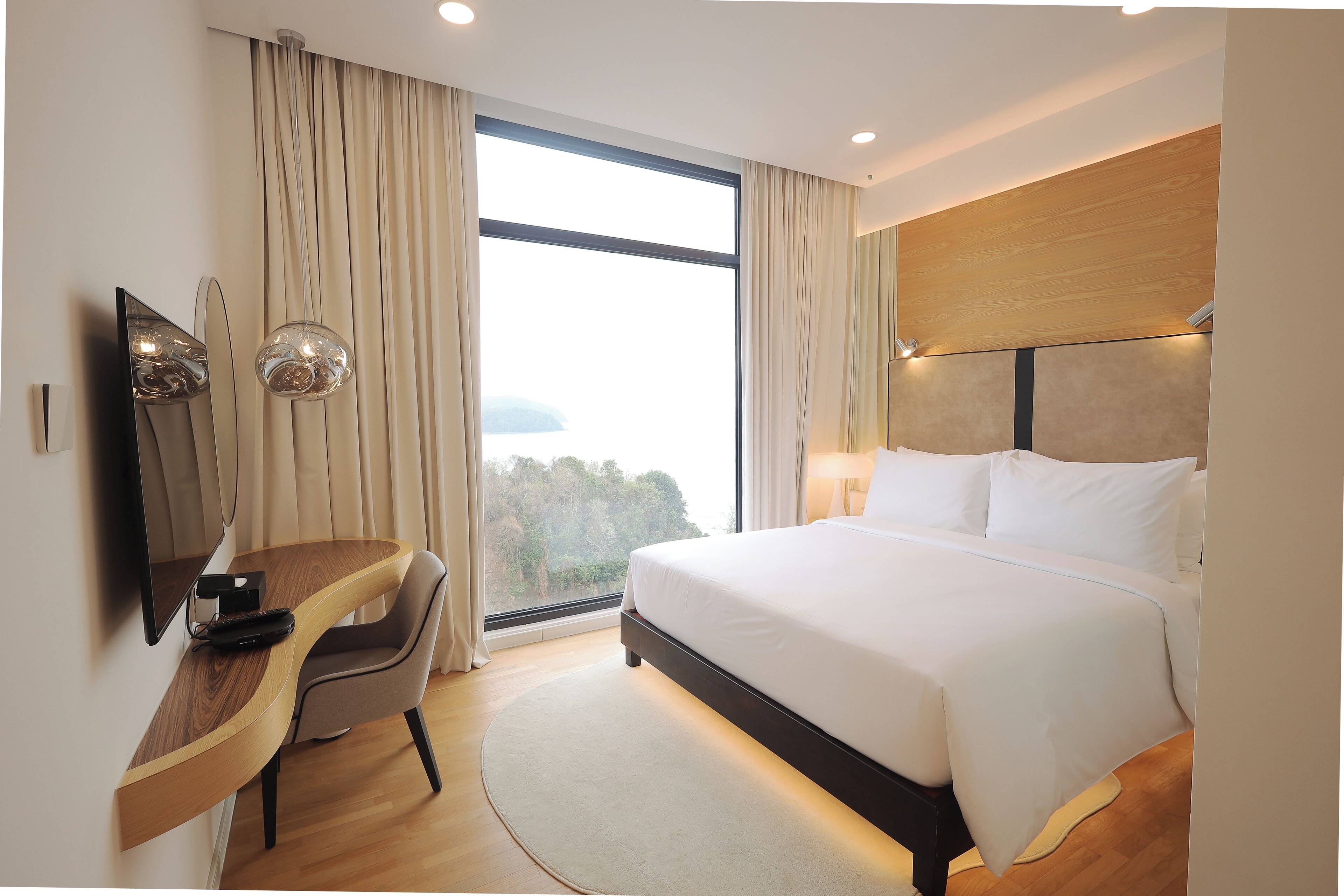 A modern hotel room with a large bed, a desk with a chair, a TV, and a large window offering a view of the outdoors.
