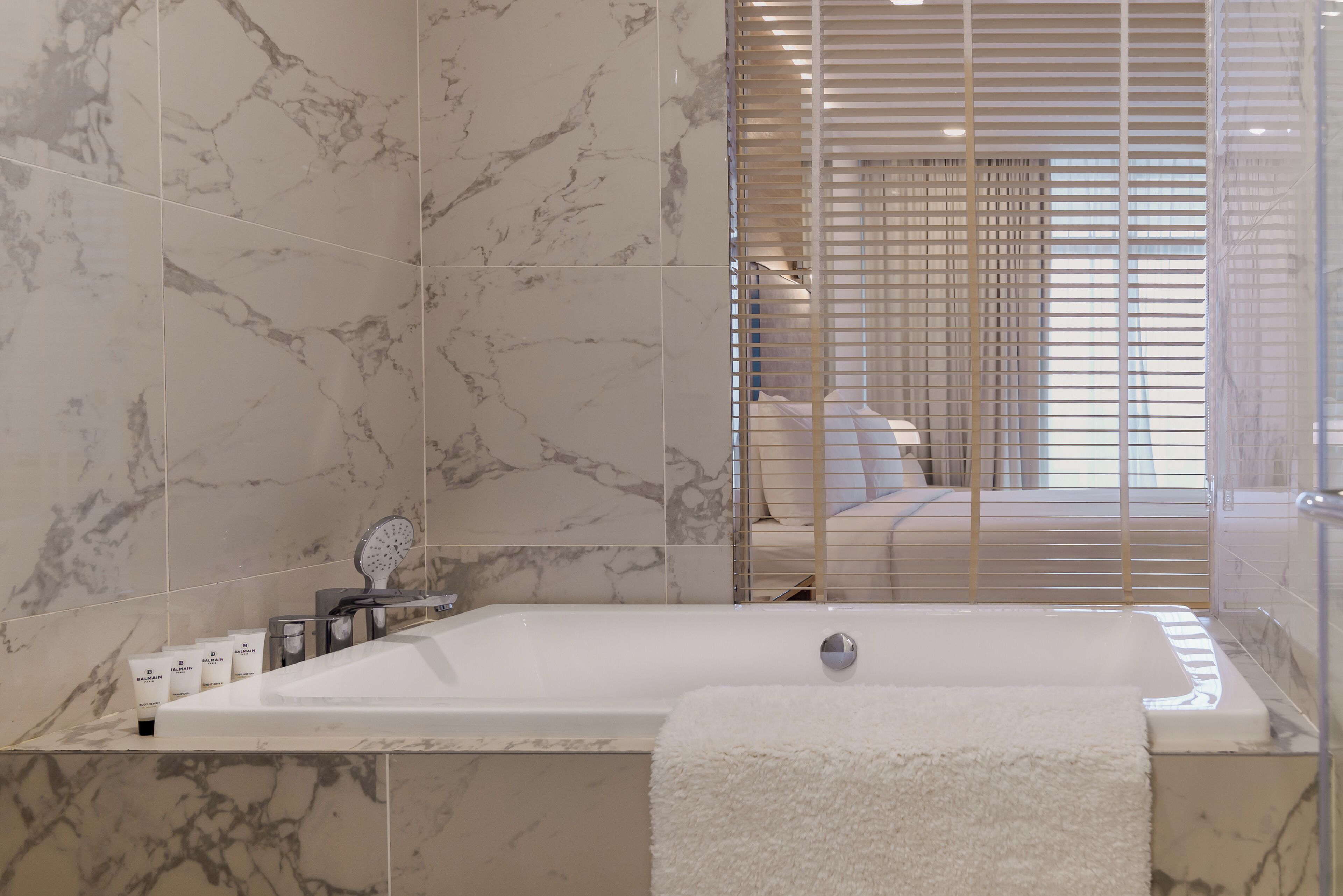 A modern bathroom with a large bathtub, marble tiles, a shower area, and a visible bed with a window in the background.