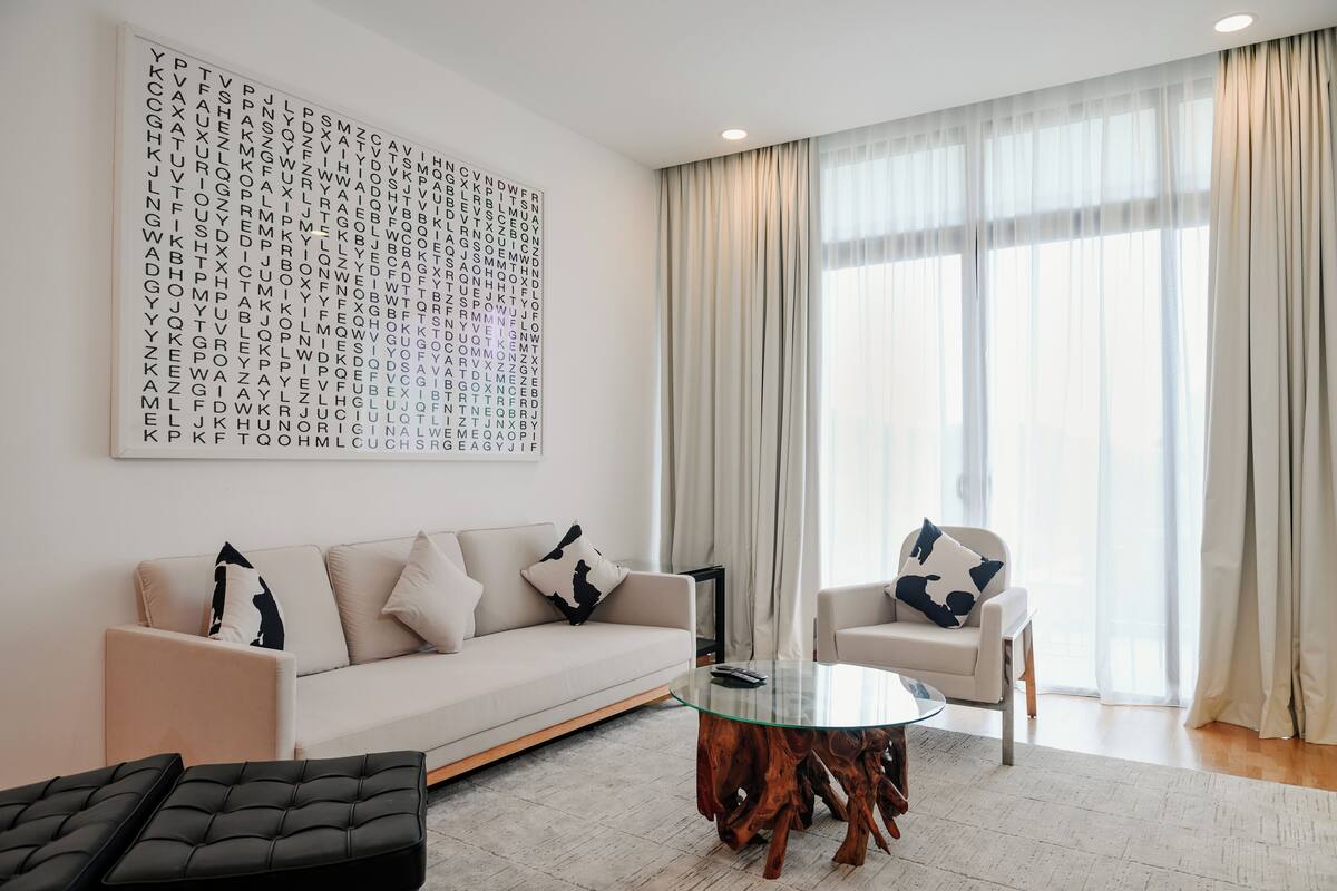A modern living room with a sofa, armchairs, a glass coffee table, and a large alphabet wall art piece.