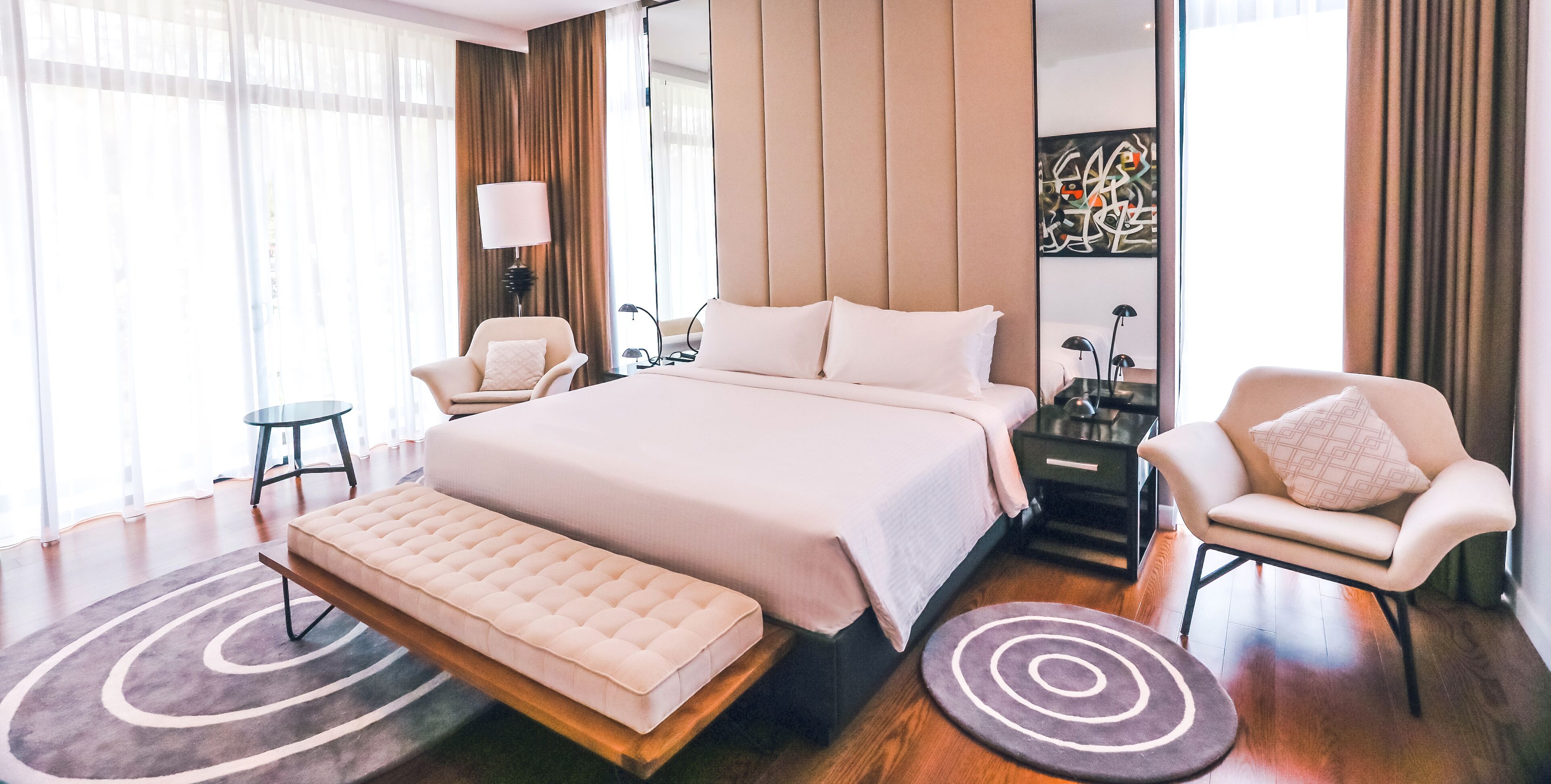 Quarto de hotel moderno com uma cama grande, duas poltronas, uma mesinha e uma mesa de cabeceira com abajur.